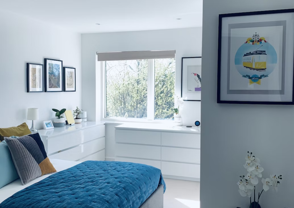 A modern bedroom with a blue and white color scheme, featuring a large window, framed artwork, and minimalistic furniture.