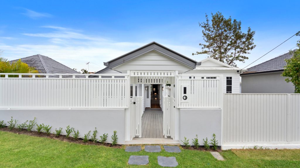 White house with a modern fence and gate, slate path, and green lawn in front. The sky is clear and blue.