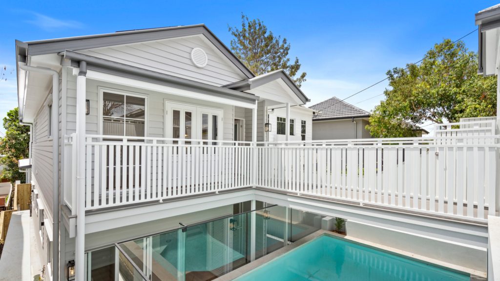 A two-story house with white siding and a wooden balcony overlooks a swimming pool. The sky is clear and blue.