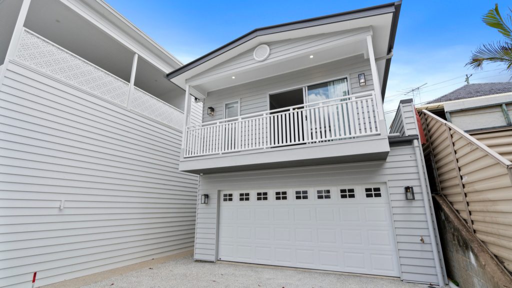 Two-story white house with a garage. Upper balcony features a railing, and the exterior is clad in horizontal siding. Blue sky in the background.