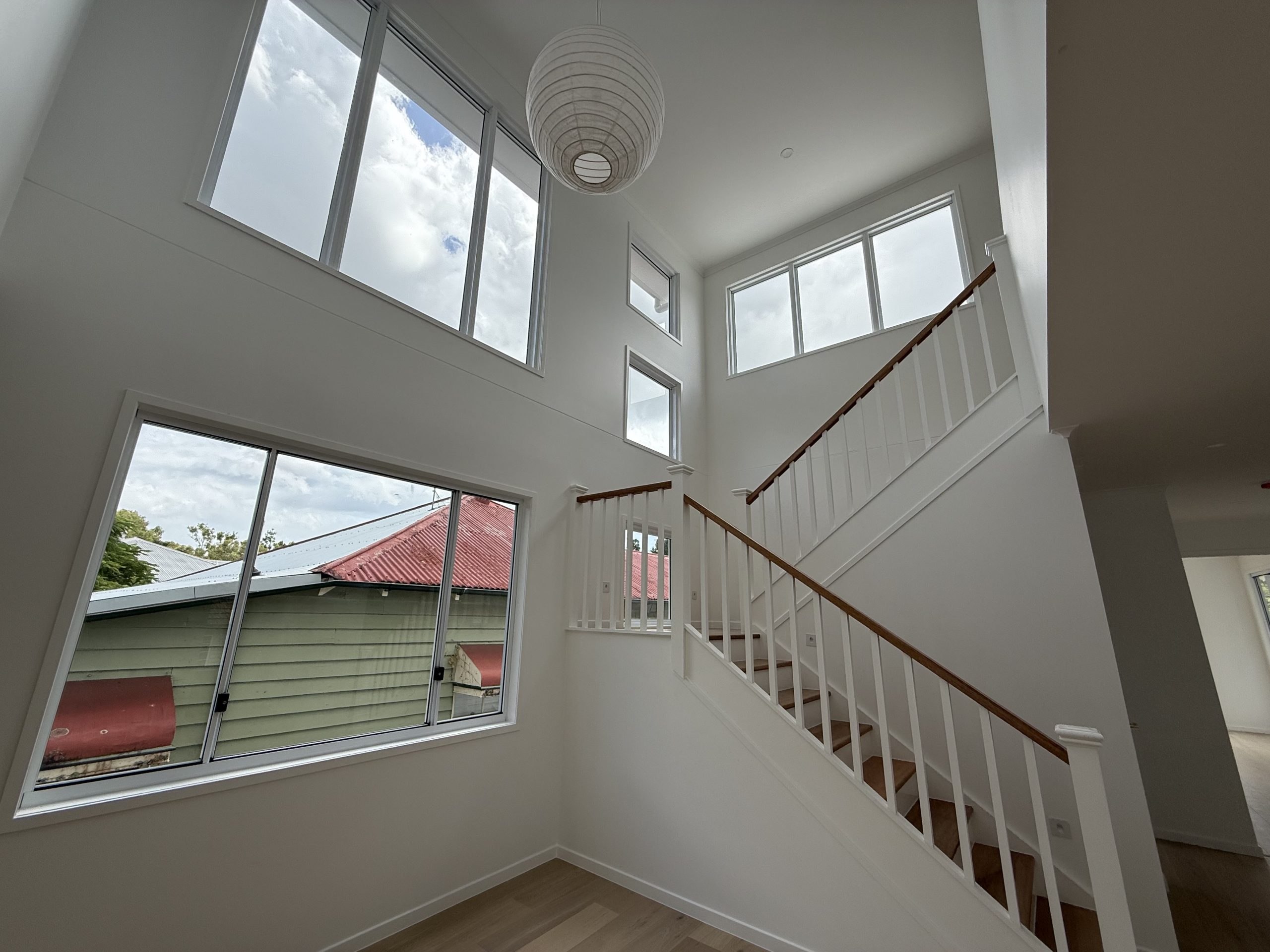 Interior of a house with large windows, high ceiling, and staircase with wooden handrail. Light fixture hangs from the ceiling.