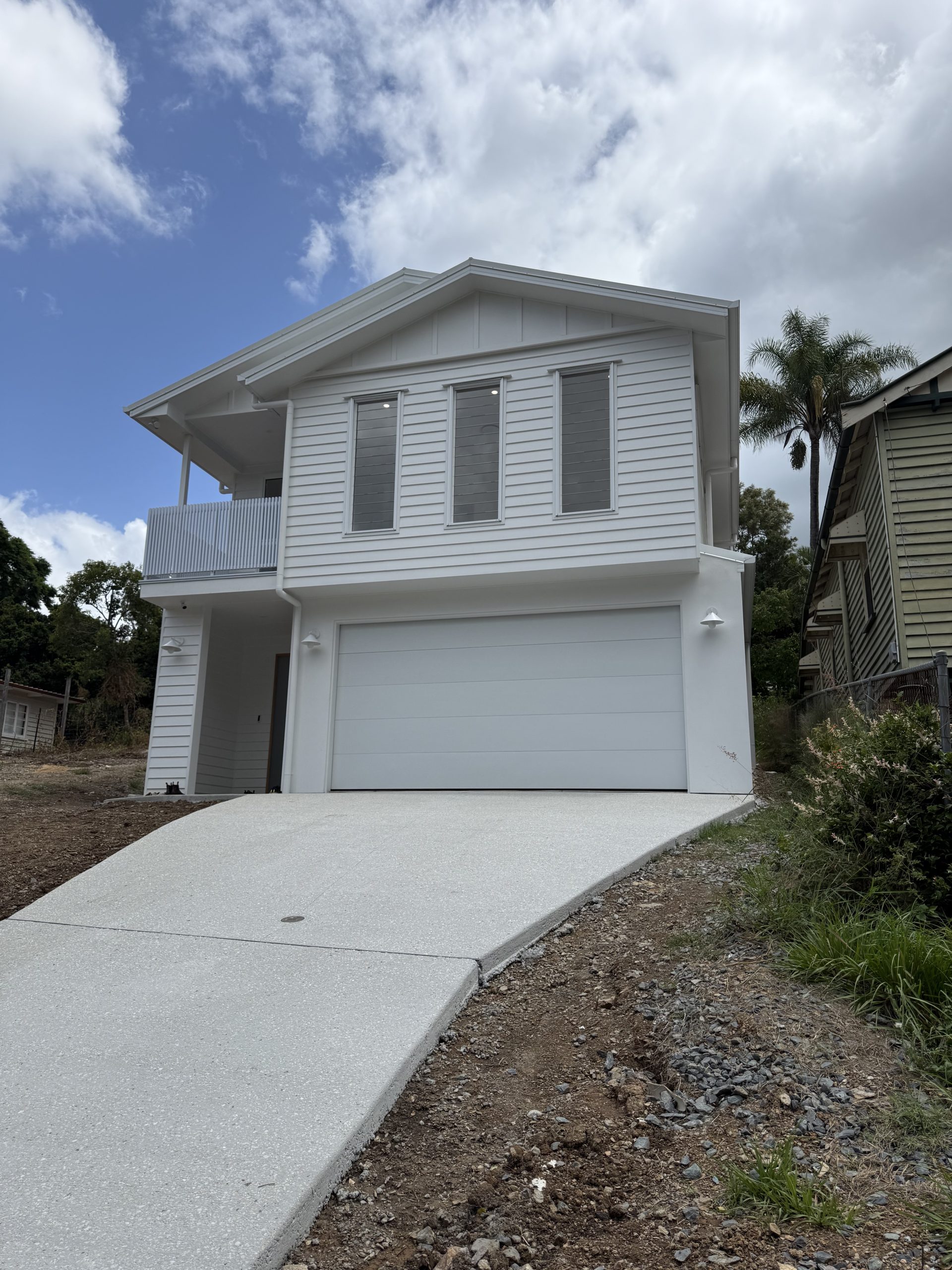 A modern, white two-story house with a garage and balcony, situated on a sloped driveway under a cloudy sky. There are trees and neighboring houses in the background.