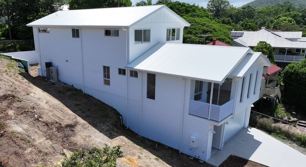 Modern white two-story house with a sloped metal roof, surrounded by a partially landscaped yard and neighboring houses in a green, hilly environment.