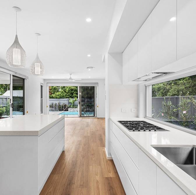 A modern kitchen in custom homes features a white island, hanging lights, gas stove, and sink. Elegant wood flooring guides you to a room with sliding glass doors that overlook a patio and sparkling swimming pool.