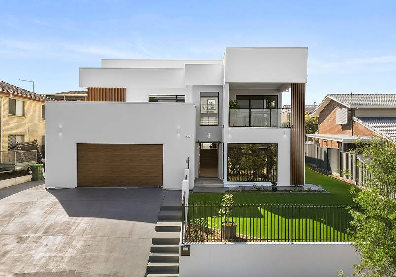 This custom home is a modern two-story white masterpiece, featuring a flat roof, large windows, and a wooden garage door. Nestled on a green lawn under the clear blue sky, it epitomizes contemporary elegance.