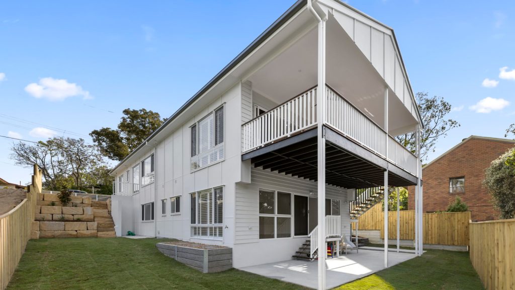 Two-story white house with a covered balcony and patio, fenced backyard, retaining wall on the left, and neatly trimmed grass.