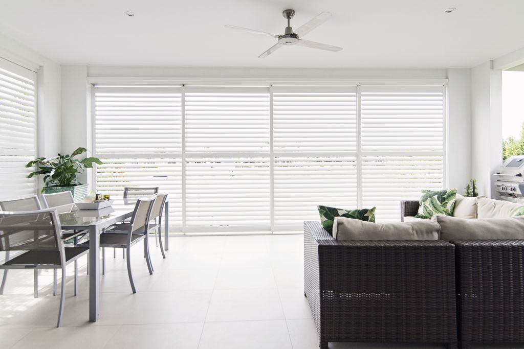 Modern sunroom with white tiled floor, glass dining table with chairs, wicker sofa with cushions, potted plants, and large windows with white blinds.